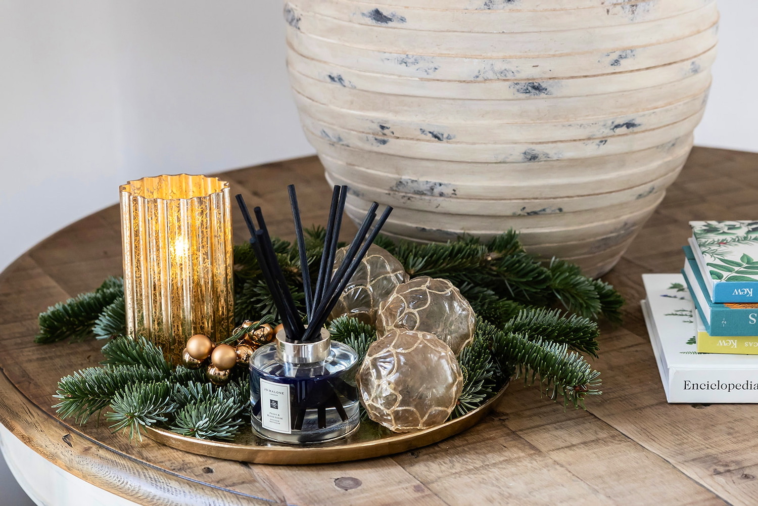 Decoración navideña sobre bandeja dorada con vela en portavelas dorado, difusor de aroma con varillas negras, esferas decorativas brillantes y ramas de pino, junto a libros apilados sobre mesa de madera rústica