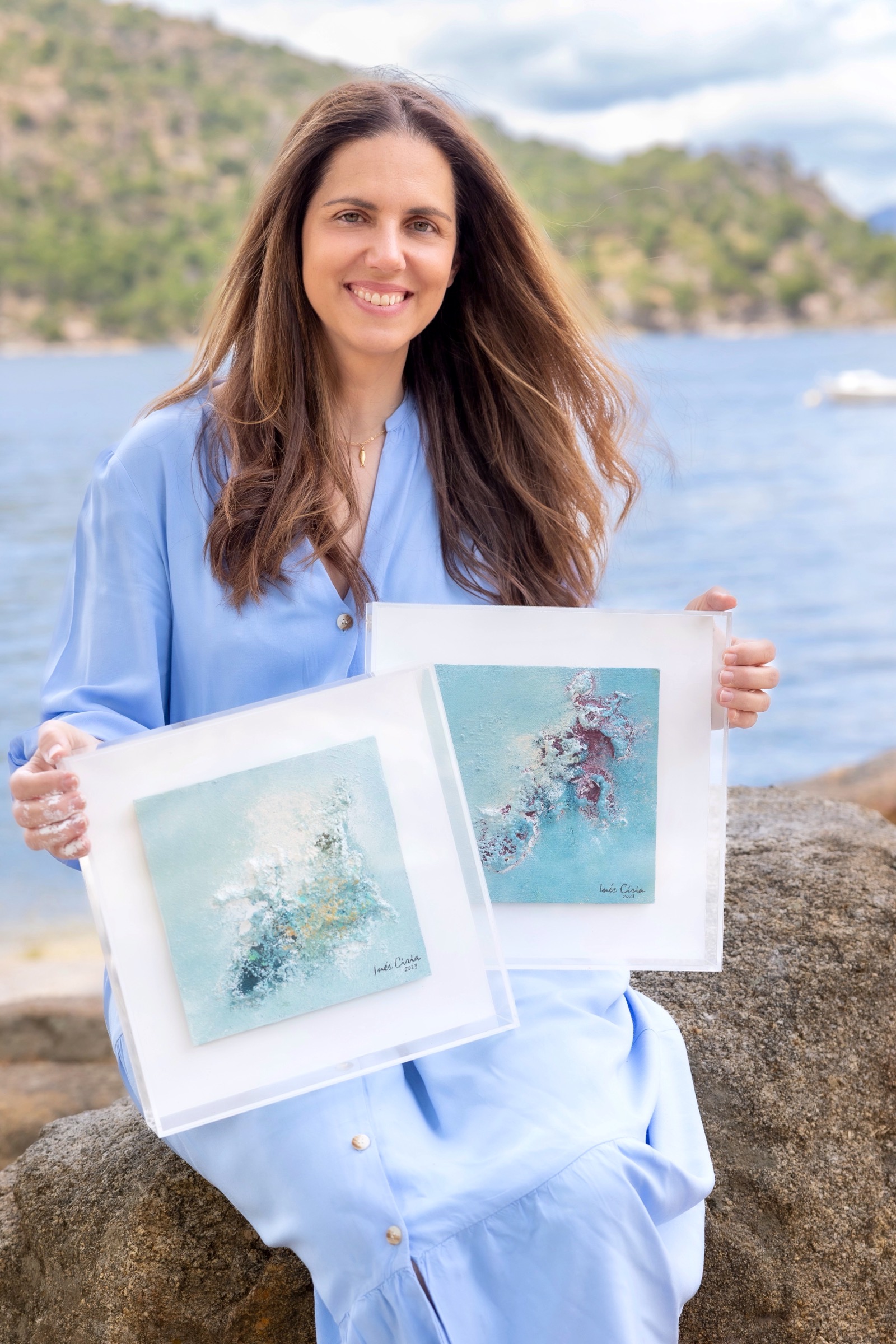 Mujer sonriente con cabello largo sosteniendo dos pinturas abstractas en tonos azules y blancos. Está sentada en una roca junto a un lago con montañas en el fondo, vistiendo una blusa azul claro.