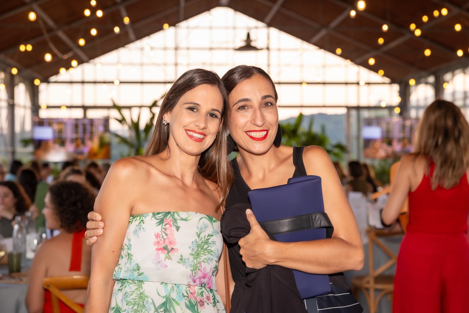 Dos mujeres elegantemente vestidas sonriendo juntas en un evento con iluminación cálida y decoración con plantas