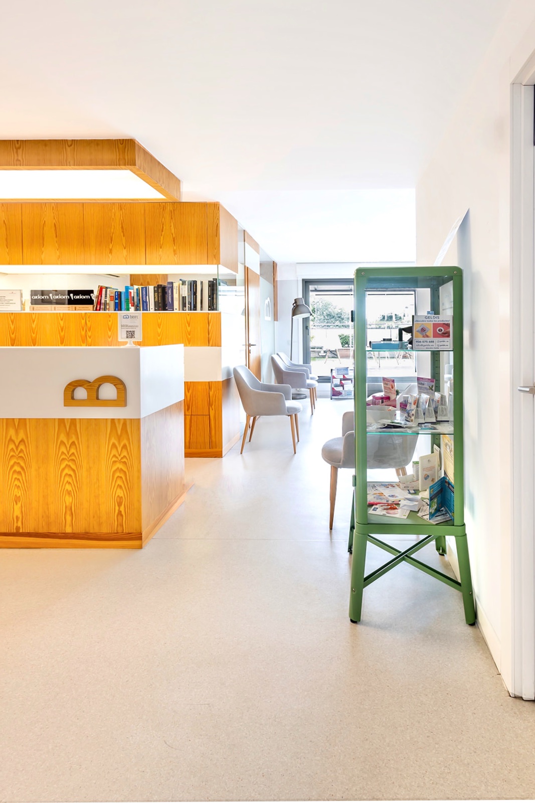 Interior moderno de un hotel con una biblioteca de madera, sillas grises y una vitrina verde. El espacio es luminoso con paredes blancas y suelo claro, creando una atmósfera acogedora y elegante.