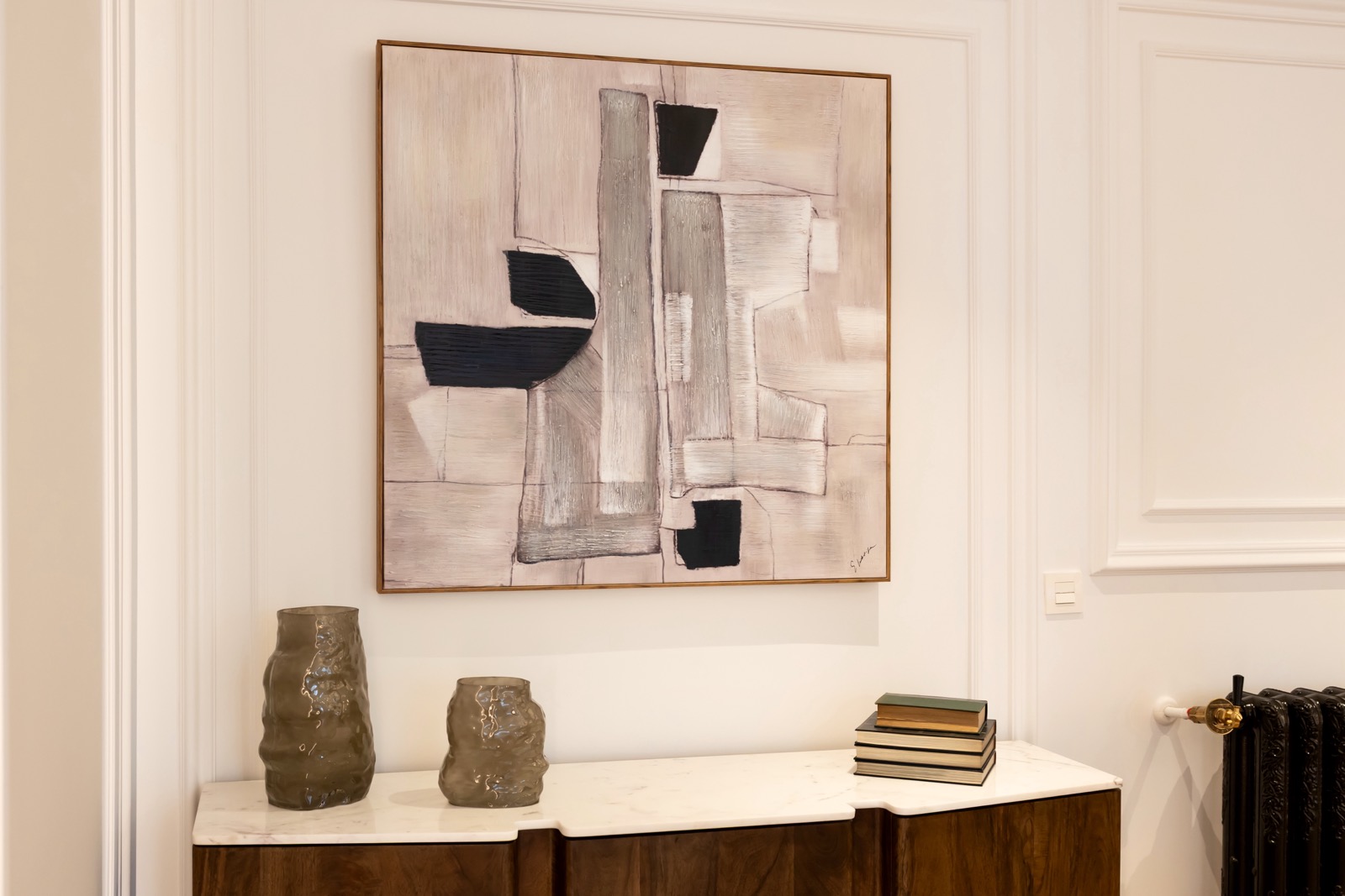 Pintura abstracta en tonos grises y negros colgada sobre un aparador de madera con superficie de mármol. Dos jarrones grises y una pila de libros adornan el aparador.