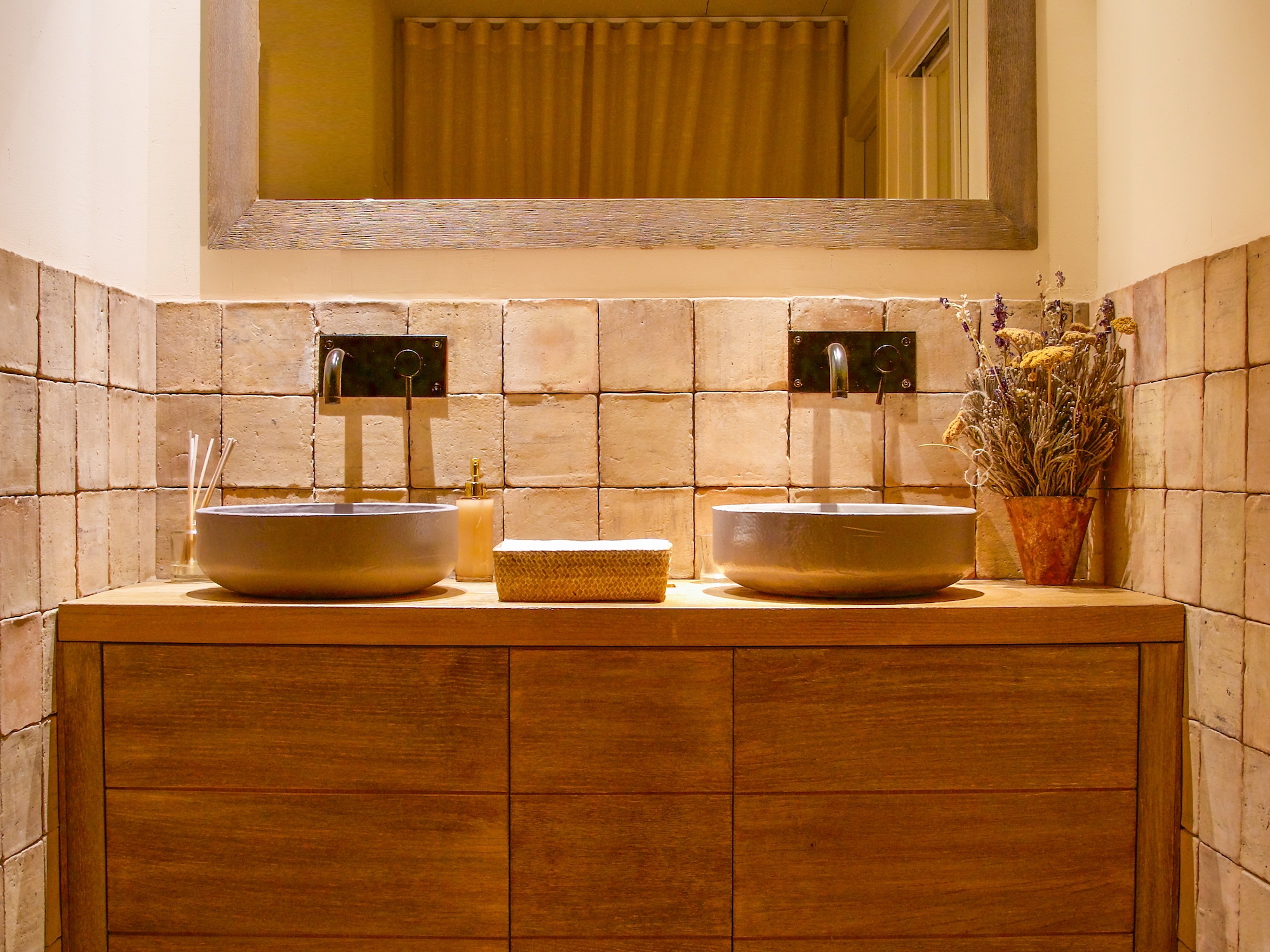Baño elegante con estilo rústico moderno que presenta un mueble de madera con dos lavabos tipo bowl, grifería negra empotrada, paredes de piedra clara y un arreglo de flores secas en maceta de barro