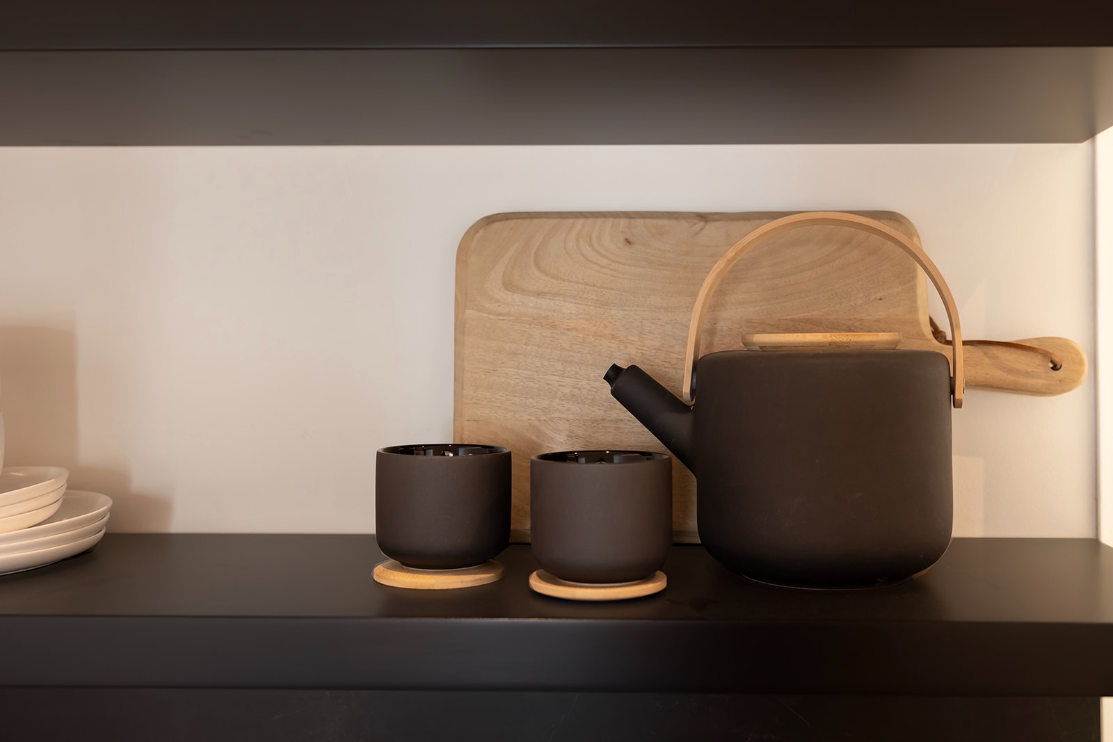 Juego de té japonés minimalista en negro y madera, que incluye una tetera y dos tazas sobre una repisa. Detrás hay una tabla de cortar de madera.