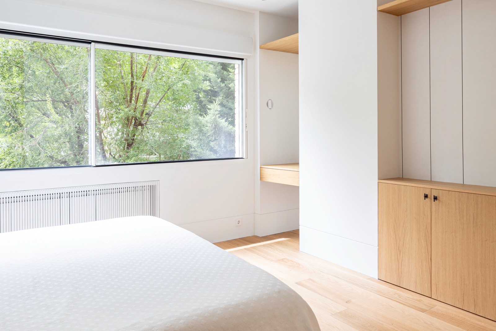 Interior de dormitorio moderno y luminoso con cama blanca, piso de madera y gran ventanal con vista a árboles verdes