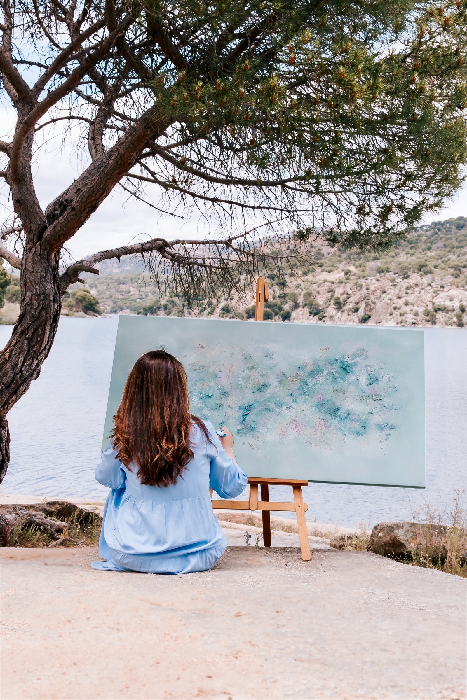 Una artista sentada de espaldas frente a un lienzo, pintando un paisaje con un lago y montañas. Está bajo un árbol con el lago y las colinas de fondo.