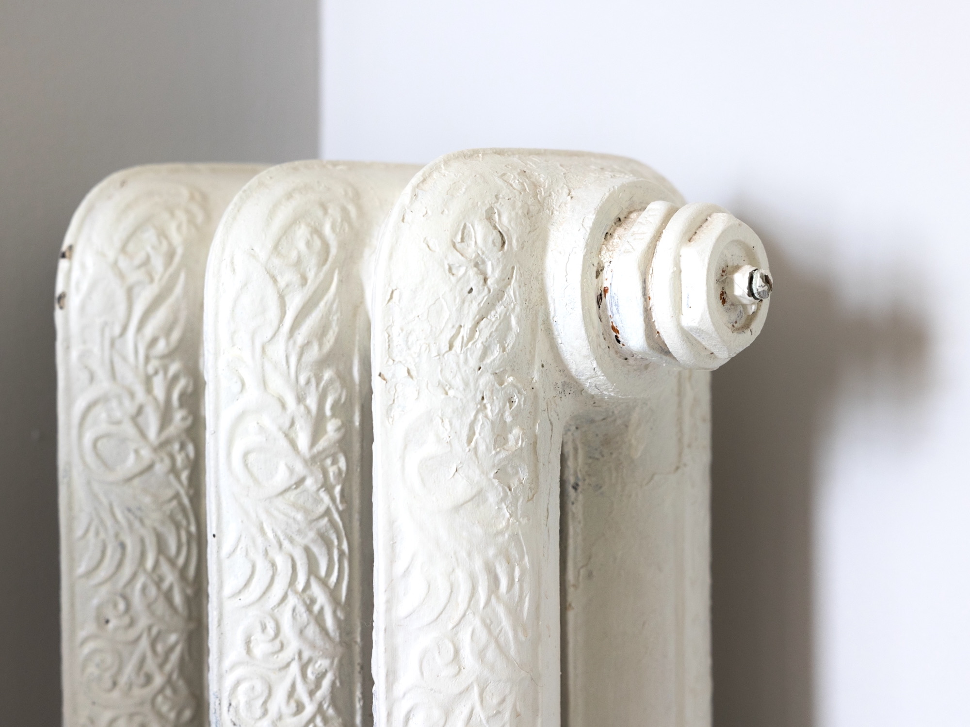 Detalle de un radiador antiguo de hierro fundido pintado en blanco con elaborados motivos florales en relieve. Se aprecian tres secciones y una válvula terminal circular en la parte superior.