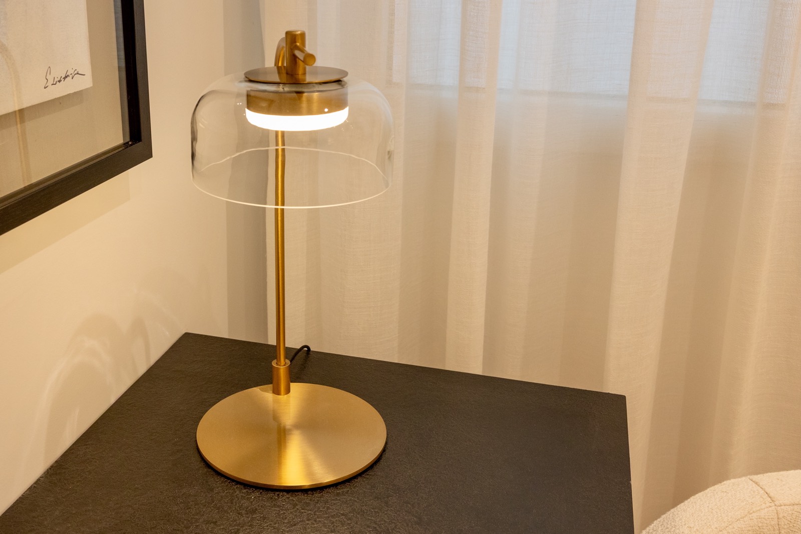 Lámpara de mesa moderna con base dorada y pantalla de cristal transparente, colocada sobre una superficie oscura. La lámpara tiene un diseño elegante y minimalista, con un aro de luz LED en la parte superior.