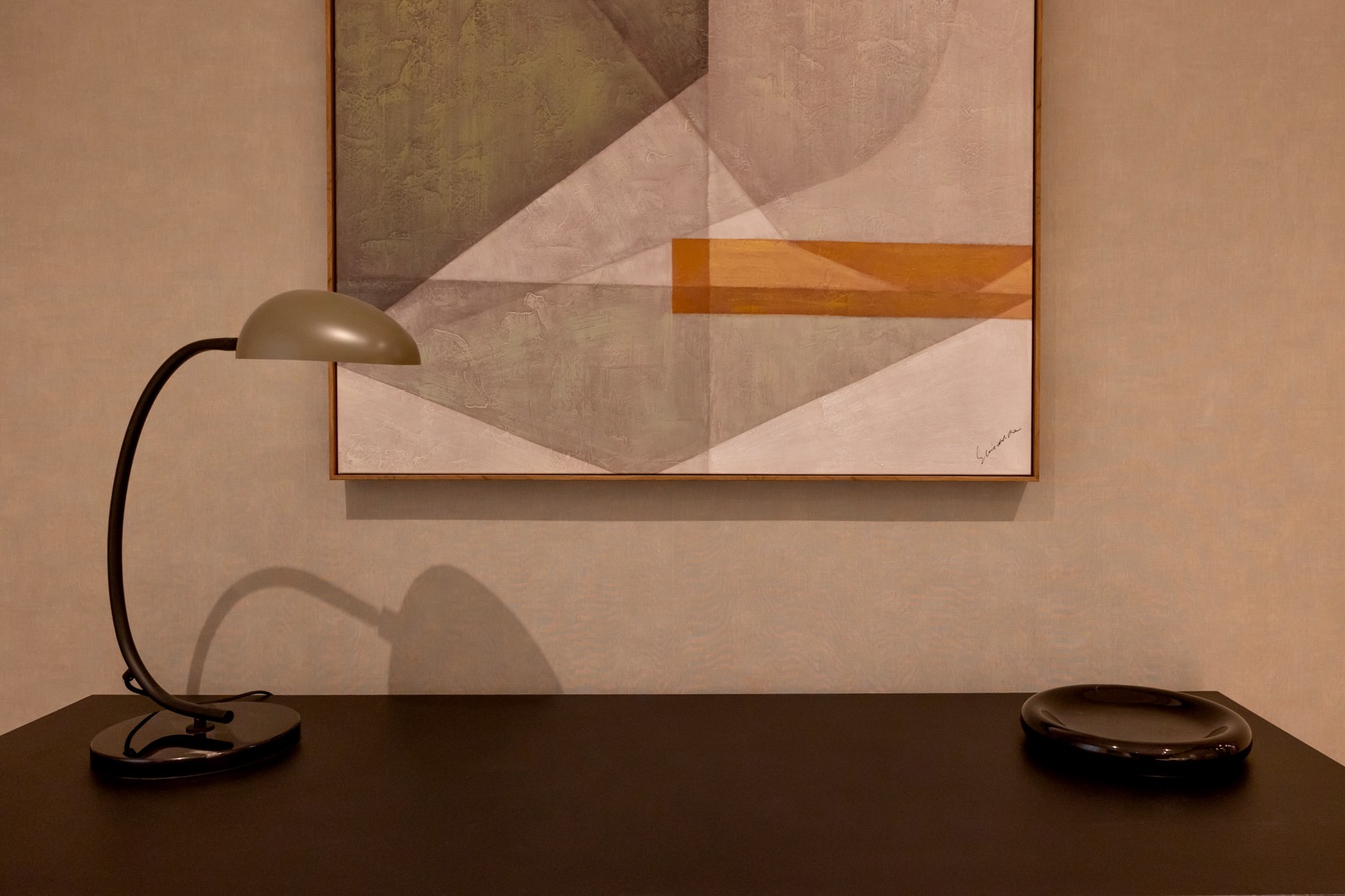 Lámpara de escritorio estilo moderno sobre mesa oscura frente a pintura abstracta geométrica en tonos pastel colgada en pared beige