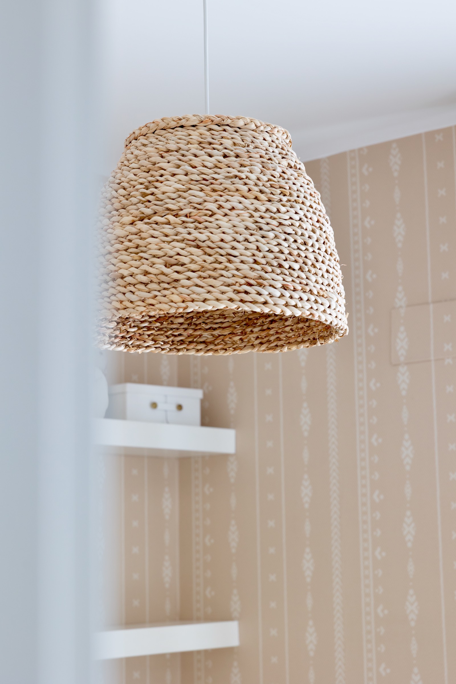 Pantalla de lámpara artesanal tejida en tonos beige y marrón, con textura de mimbre trenzado, suspendida frente a una pared clara