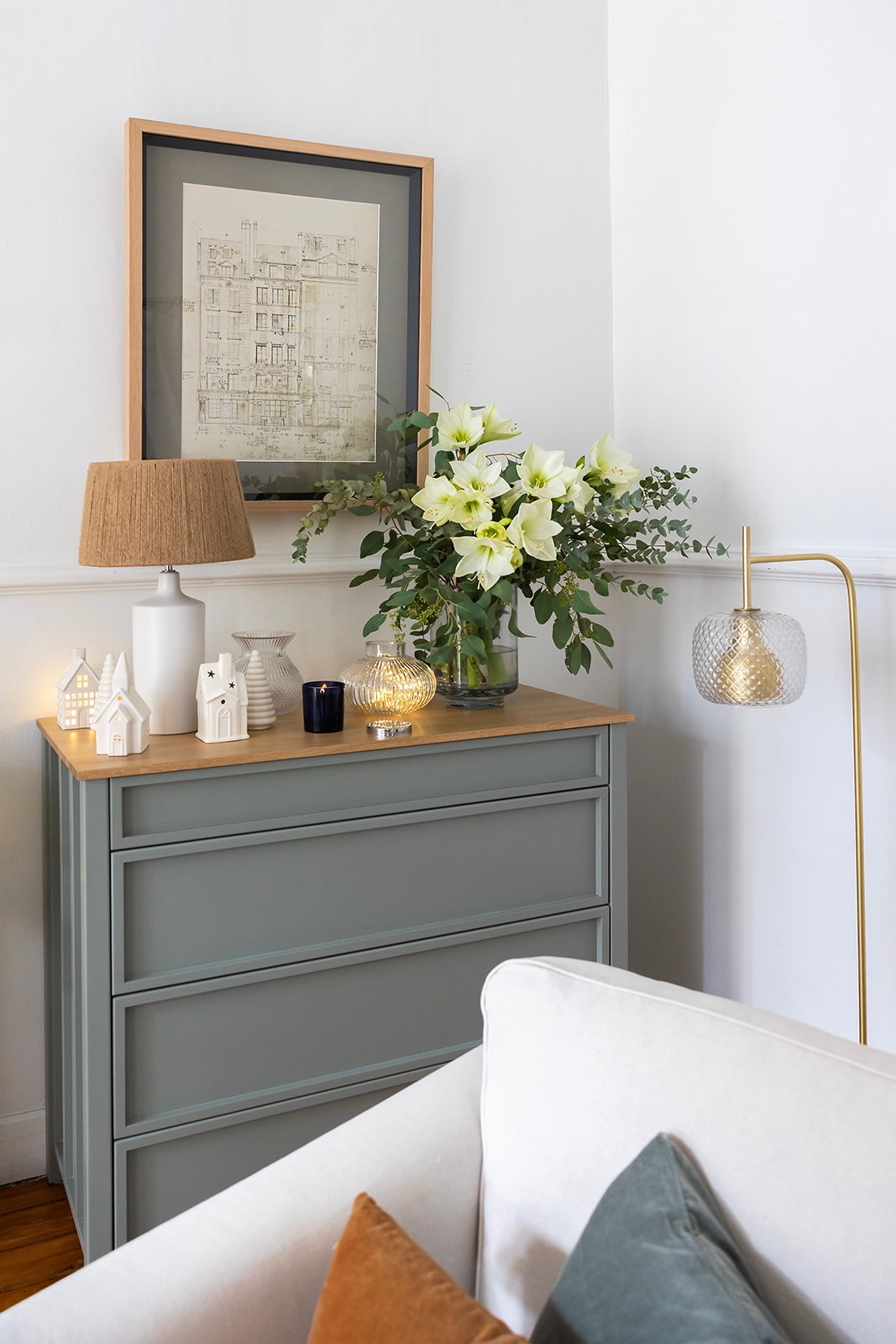 Cómoda gris con superficie de madera decorada con flores amarillas, lámpara, casitas decorativas y velas. Sobre ella, un cuadro con plano arquitectónico enmarcado. Junto a la cómoda, una lámpara de pie dorada y un sofá blanco.