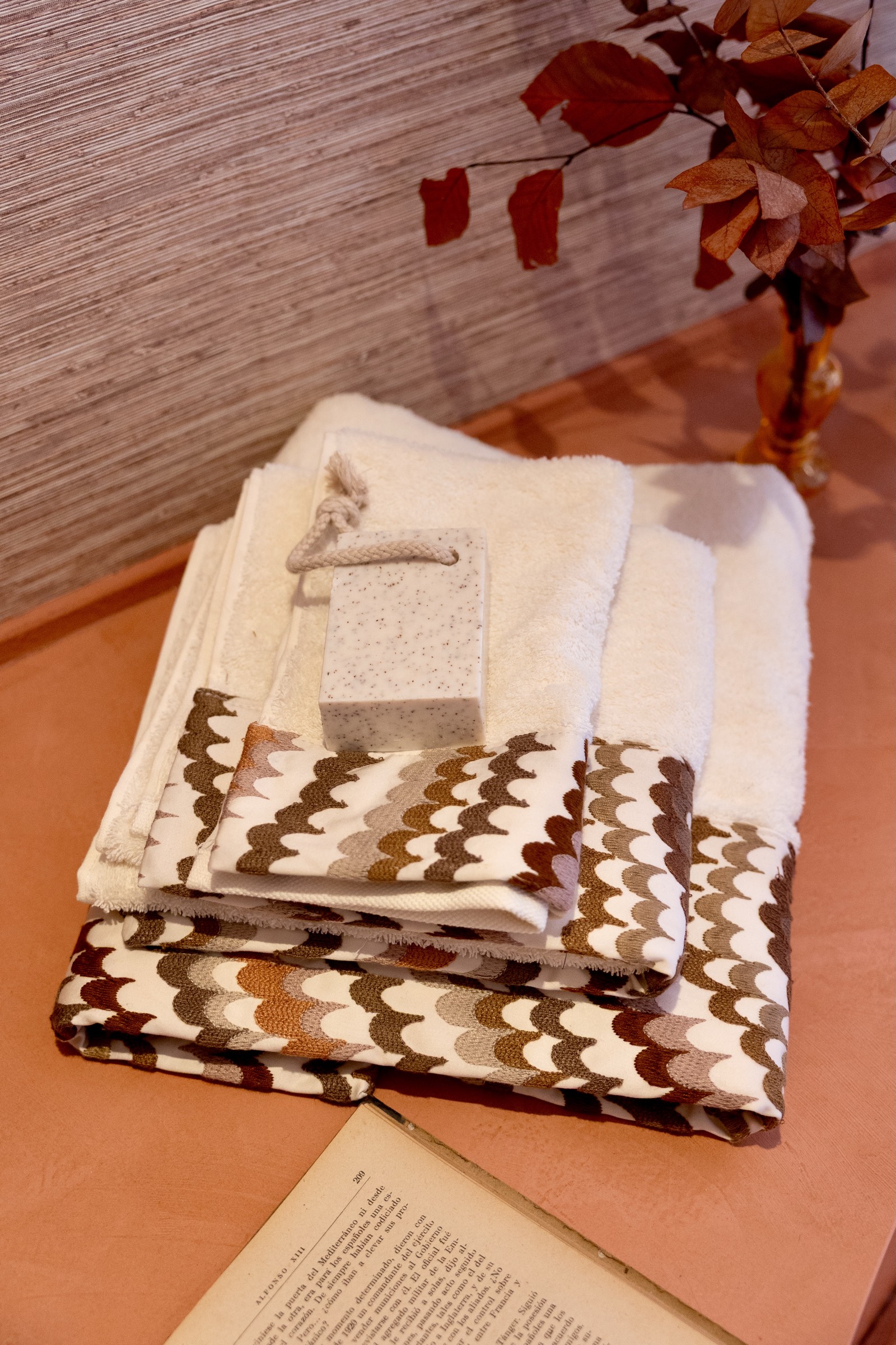 Toallas blancas dobladas con un patrón ondulado en tonos marrones, junto a un jabón de piedra y una rama con hojas rojas y naranjas