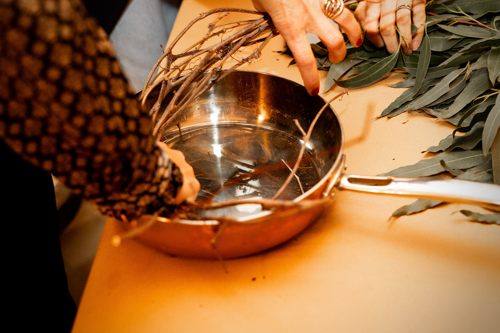 Manos preparando una infusión de hierbas en un recipiente metálico. Se observan ramas y hojas frescas sobre una mesa, junto a un cuenco brillante donde se mezclan los ingredientes.