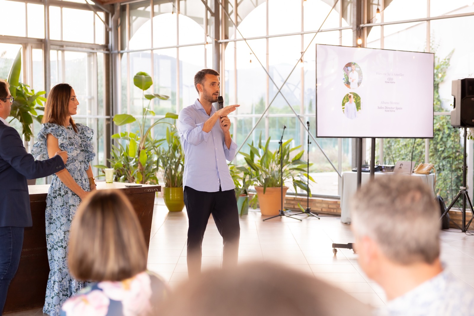 Presentación empresarial en una luminosa sala de reuniones con plantas verdes, un presentador de pie y una pantalla con diapositivas