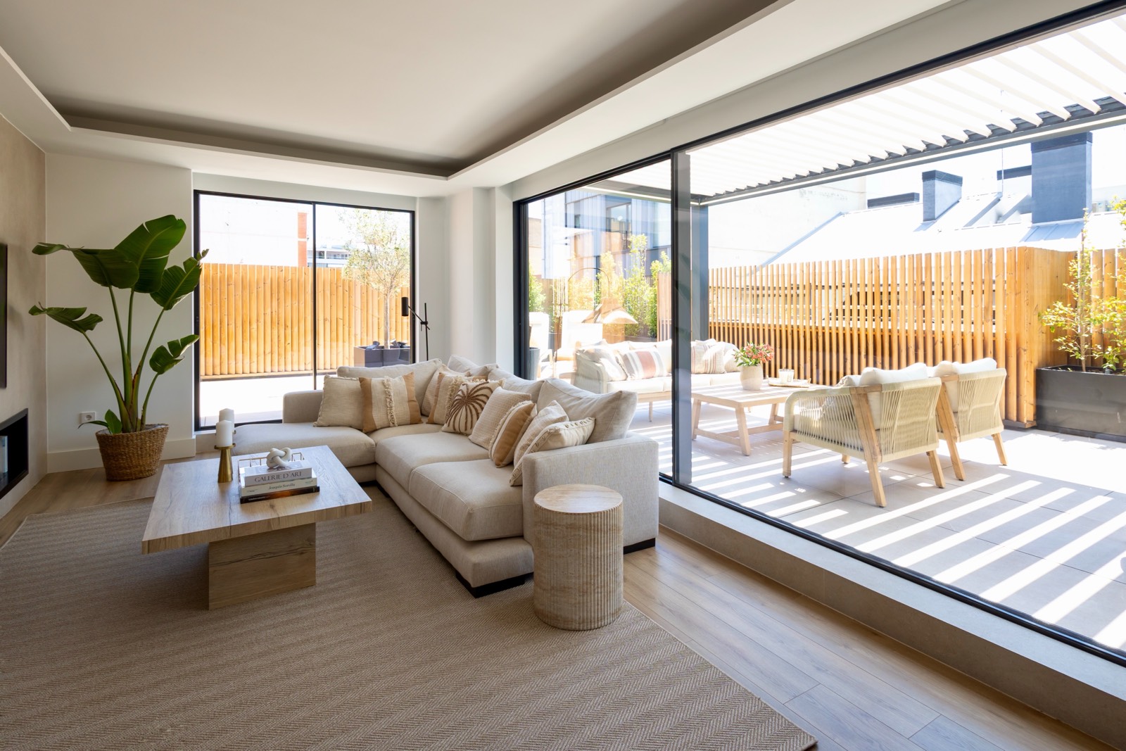 Sala de estar moderna con grandes ventanales que dan a una terraza exterior. El interior tiene un sofá beige, una mesa de centro de madera y una planta grande. La terraza tiene muebles de exterior y una valla de madera.