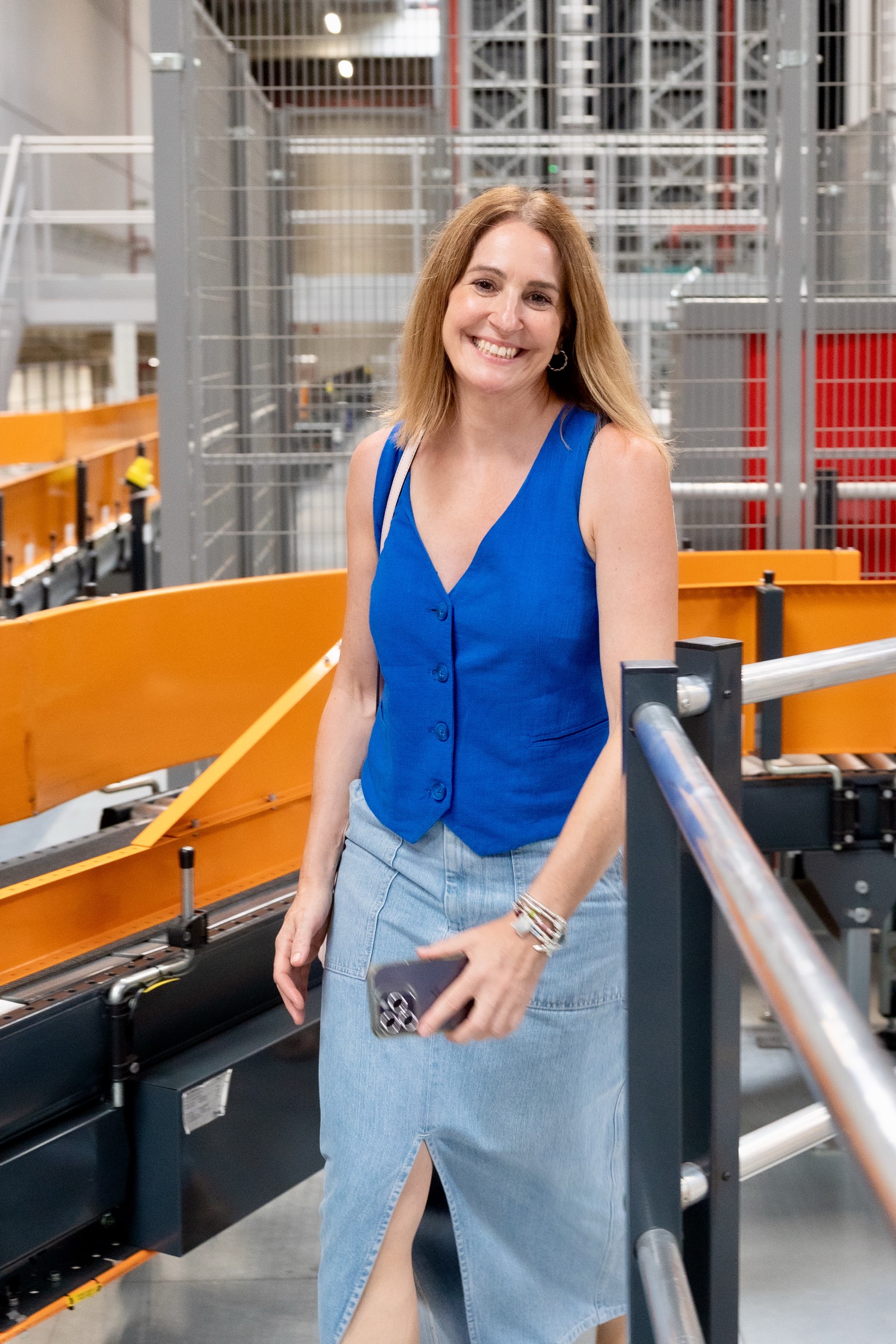 Mujer sonriente con chaleco azul y falda vaquera, de pie en un entorno industrial con máquinas y estructuras naranjas y grises
