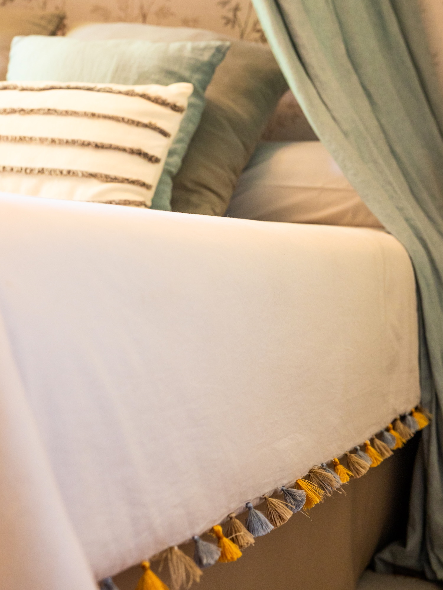 Detalle de una cama con sábanas blancas decorada con borlas amarillas y grises en el borde, almohadas decorativas con rayas y cortinas verdes suaves que crean un ambiente acogedor