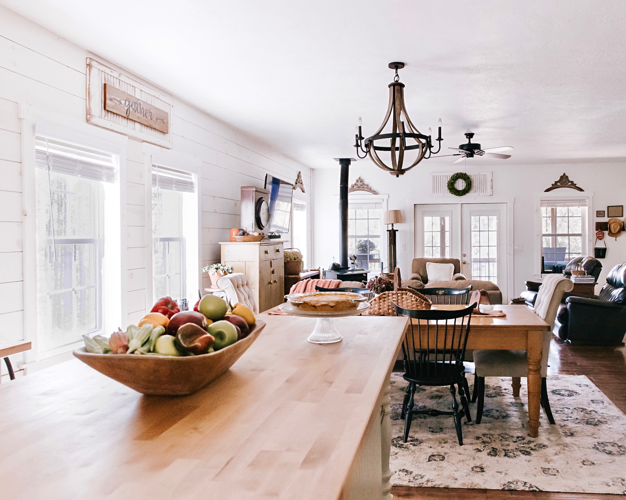 Comedor espacioso con estilo farmhouse, mesa de madera con frutero, lámpara de araña rústica, paredes blancas con revestimiento de madera, ventanas grandes y decoración campestre con letrero 'gather' en la pared