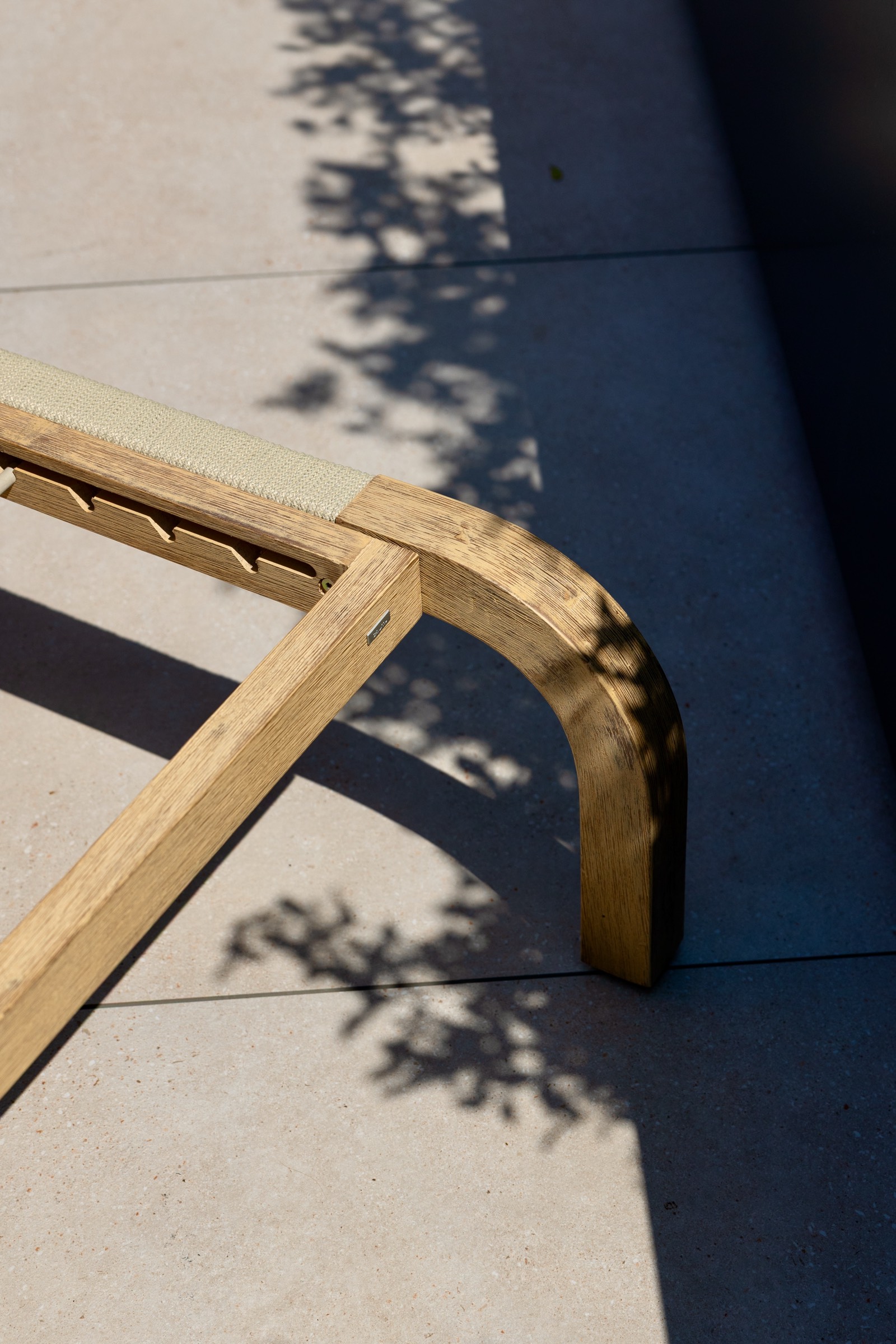 Detalle de una silla de jardín de madera con tejido beige, proyectando sombras en el suelo de baldosas. La luz del sol crea un contraste entre las áreas iluminadas y sombreadas.