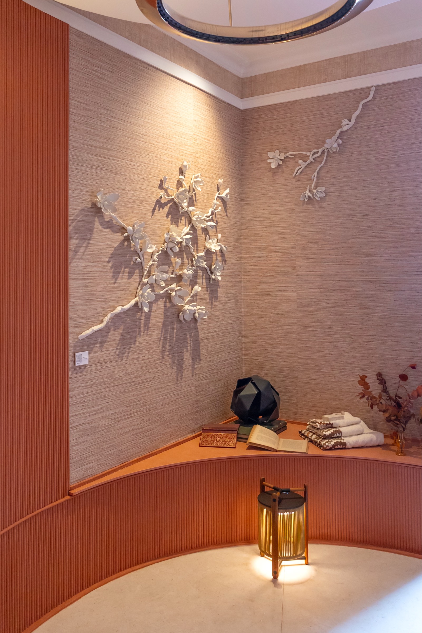 Rincón interior elegante con escultura de pared de flores blancas, lámpara dorada, toallas y decoración minimalista en tonos cálidos