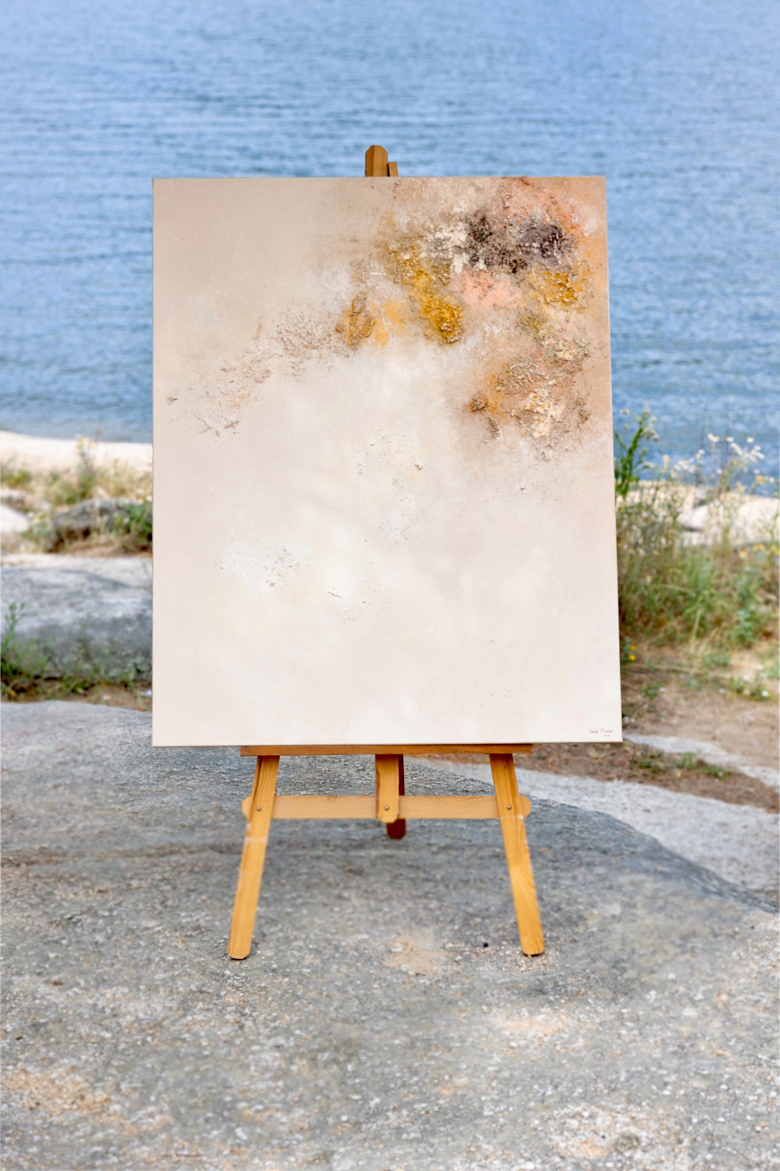 Un lienzo en blanco con manchas abstractas en la parte superior, colocado en un caballete de madera junto a un cuerpo de agua azul. El lienzo está situado en una orilla rocosa con algunas plantas.