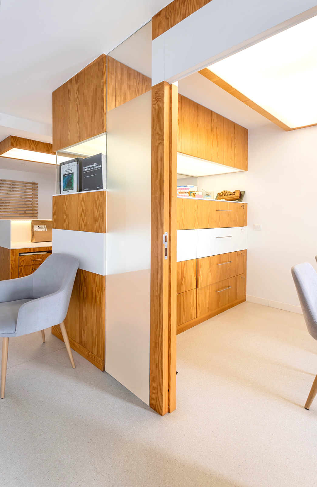 Interior moderno con muebles de madera, paredes blancas y sillas grises. Diseño minimalista con elementos de almacenamiento integrados y iluminación empotrada en el techo.