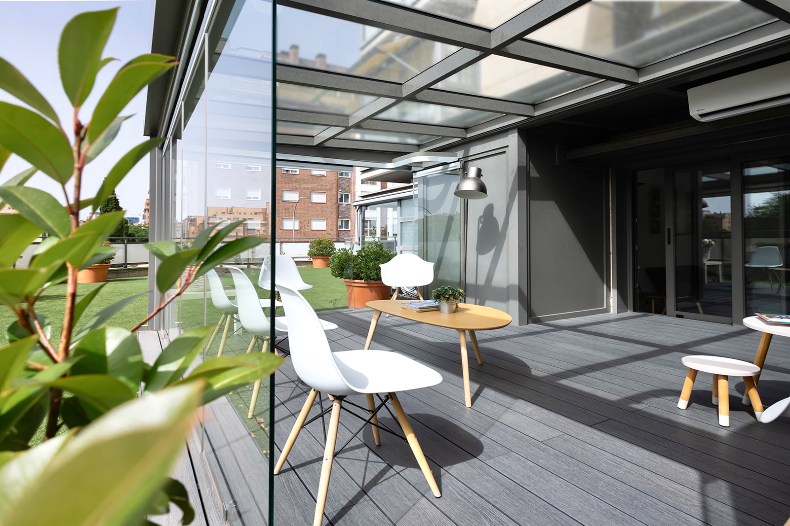 Terraza moderna acristalada con techo transparente, muebles blancos y de madera, y plantas. El espacio ofrece una vista de edificios cercanos y está decorado con un estilo minimalista y elegante.