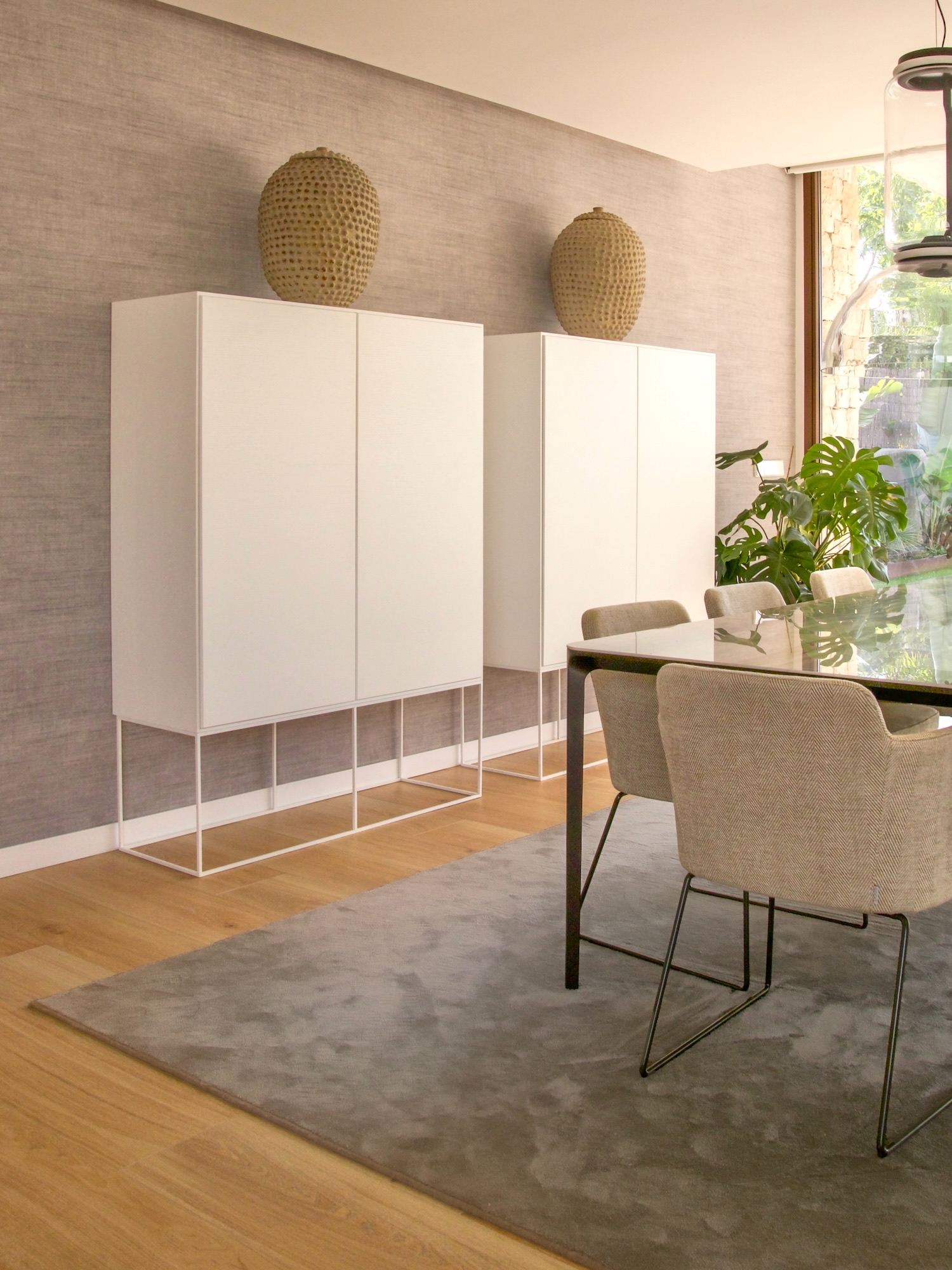 Comedor moderno y minimalista con gabinetes blancos elevados sobre bases transparentes, decorados con vasijas texturizadas, sillas tapizadas en gris y una planta monstera junto a una ventana