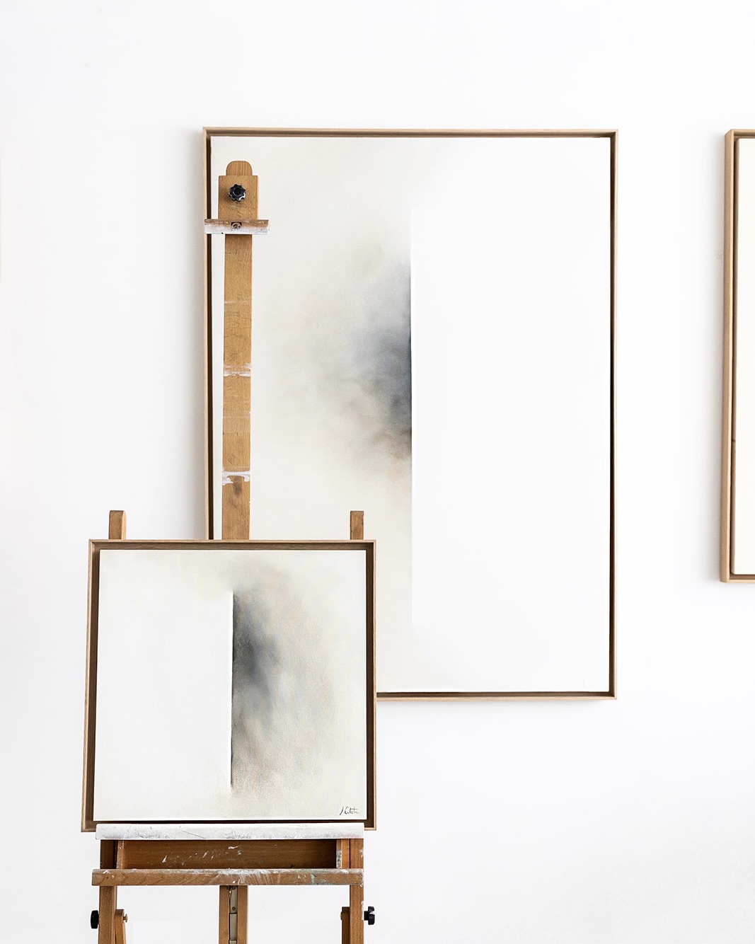 Dos lienzos con diseños minimalistas en tonos grises y blancos, uno grande en la pared y otro más pequeño en un caballete de madera. Los marcos son de madera clara y el fondo es blanco.