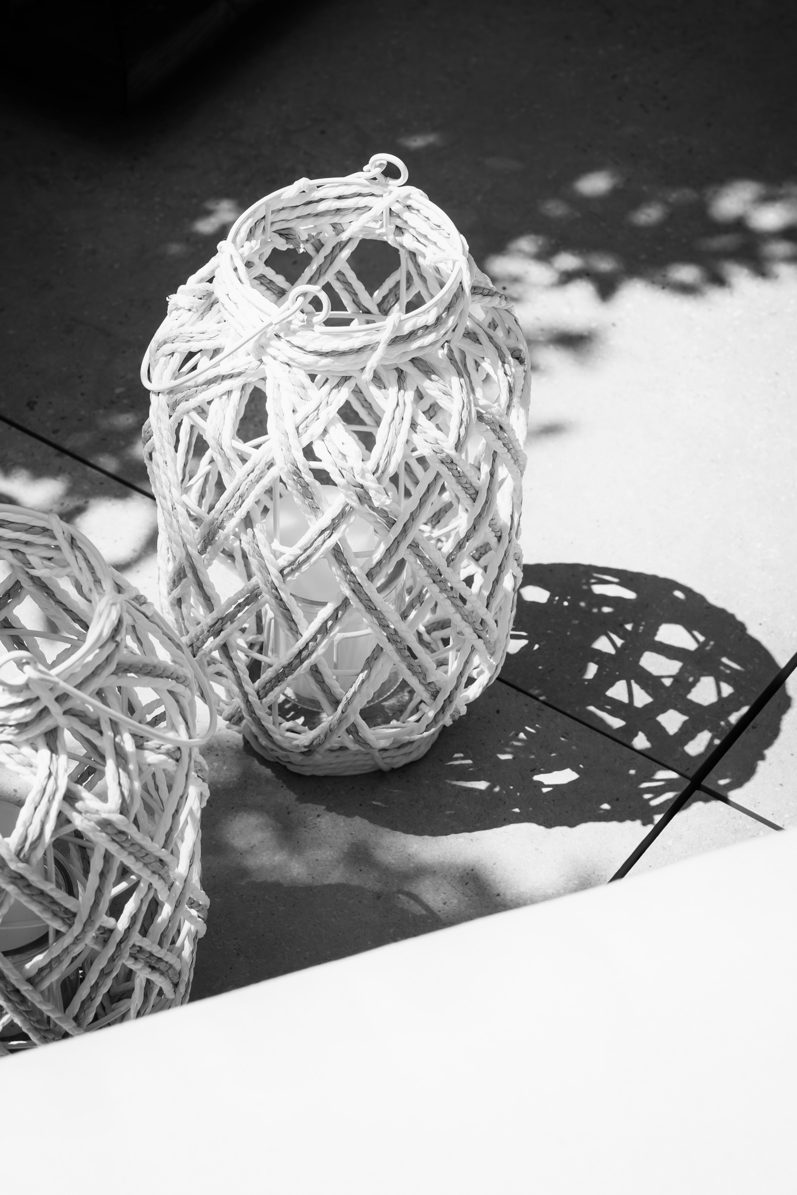 Imagen en blanco y negro de un farol decorativo tejido con fibras blancas, creando un patrón intrincado de líneas entrecruzadas. El farol proyecta una sombra detallada sobre una superficie plana.