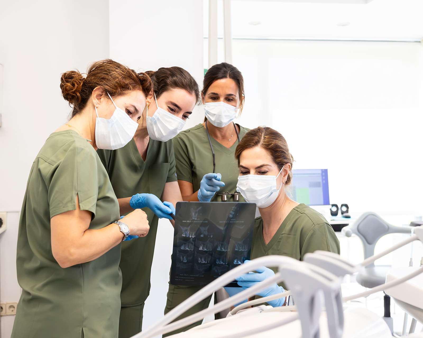 Equipo médico en uniformes verdes y mascarillas examinando una radiografía dental en una clínica. Cuatro profesionales de la salud observan atentamente la imagen en un ambiente clínico con equipos médicos visibles.