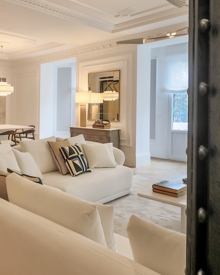 Elegante sala de estar en tonos blancos con sofá amplio, techos altos decorados, lámparas colgantes y detalles en madera. La habitación tiene un estilo clásico moderno con molduras ornamentadas y una atmósfera luminosa y espaciosa.