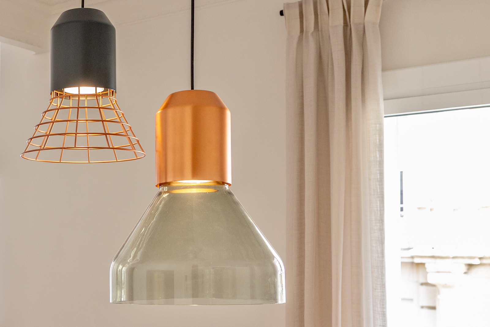 Dos lámparas colgantes modernas en una habitación. Una lámpara tiene una pantalla de vidrio transparente con base dorada, la otra tiene una rejilla metálica dorada con base negra. Las lámparas cuelgan frente a una ventana con cortinas claras.