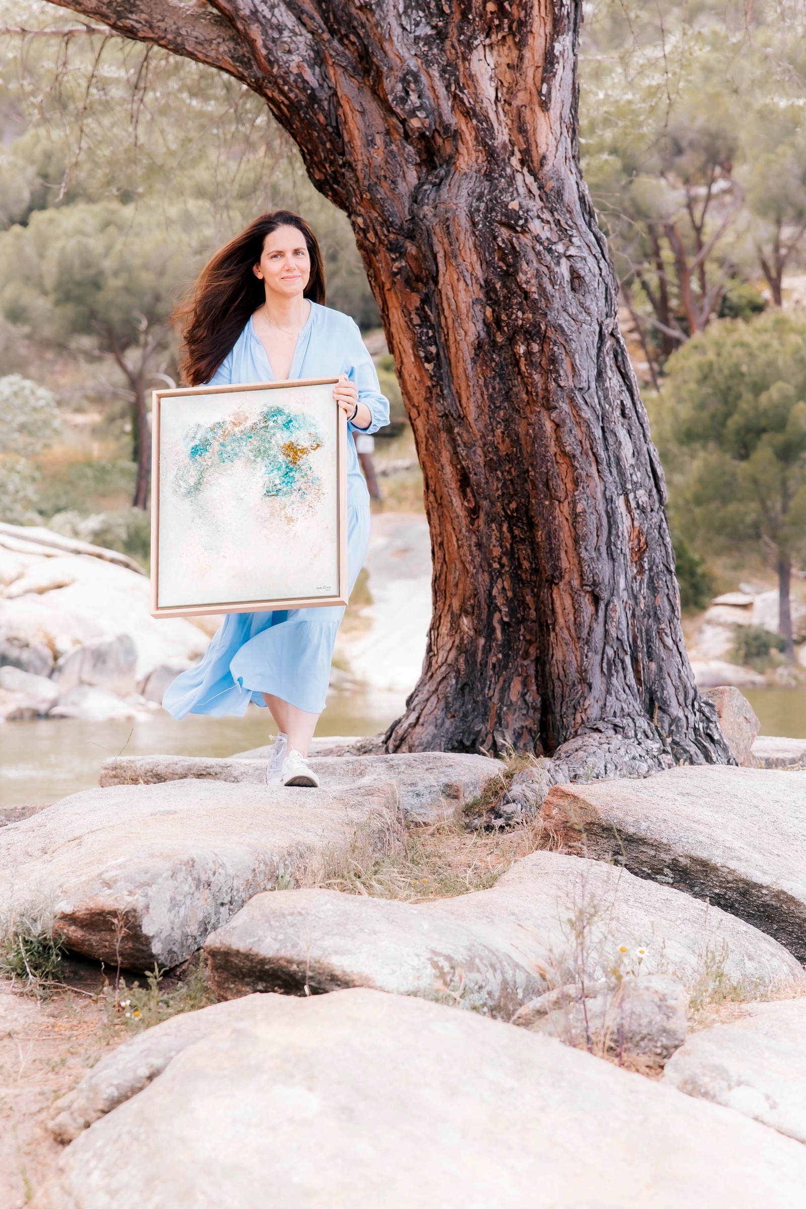 Una persona con un vestido azul sostiene un cuadro abstracto frente a un árbol antiguo en un entorno natural con rocas y vegetación