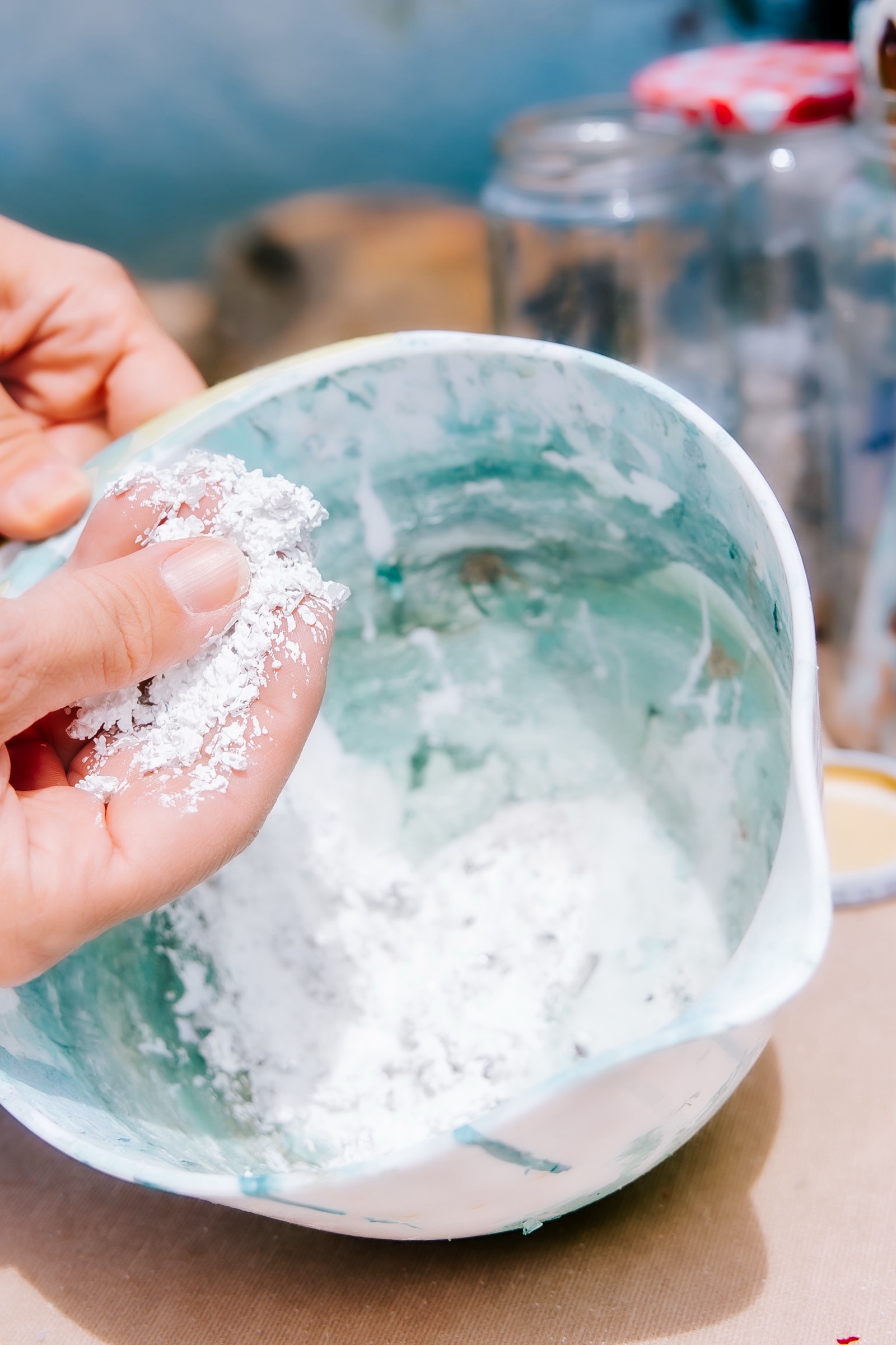 Manos amasando una mezcla blanca en un tazón de cerámica con tonos turquesa. Se observan otros recipientes de vidrio en el fondo, sugiriendo un proceso de preparación culinaria o artesanal.