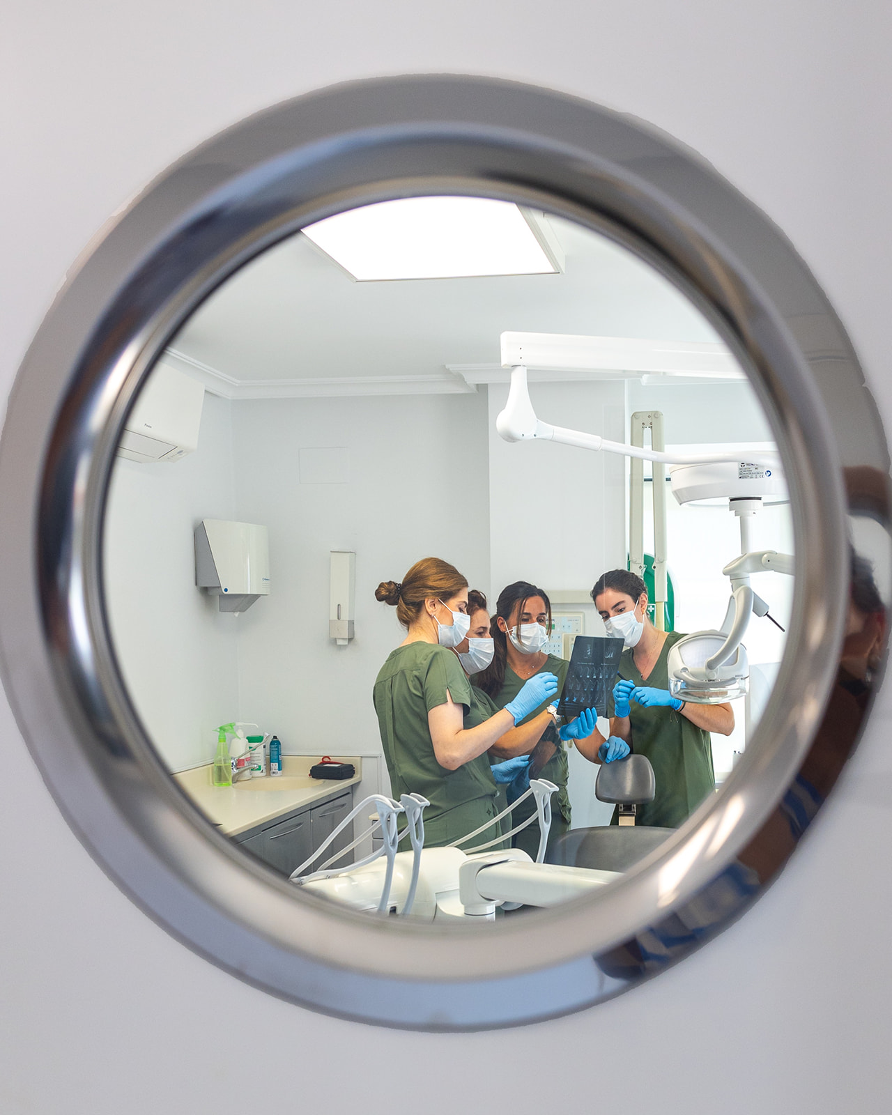 Vista a través de un espejo circular de un equipo médico en un consultorio dental. Los profesionales visten uniformes verdes y mascarillas, examinando una radiografía dental juntos.