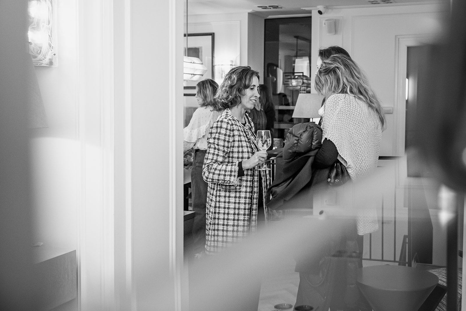 Imagen en blanco y negro de una reunión social en interiores. Dos personas conversan mientras sostienen copas de vino. La escena muestra un ambiente acogedor y elegante.