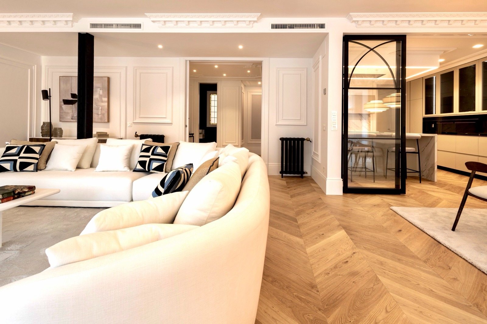 Elegante sala de estar moderna con sofá blanco, piso de madera en espiga, molduras decorativas en el techo y una puerta de cristal que separa la cocina. La decoración combina elementos clásicos y contemporáneos en tonos neutros.