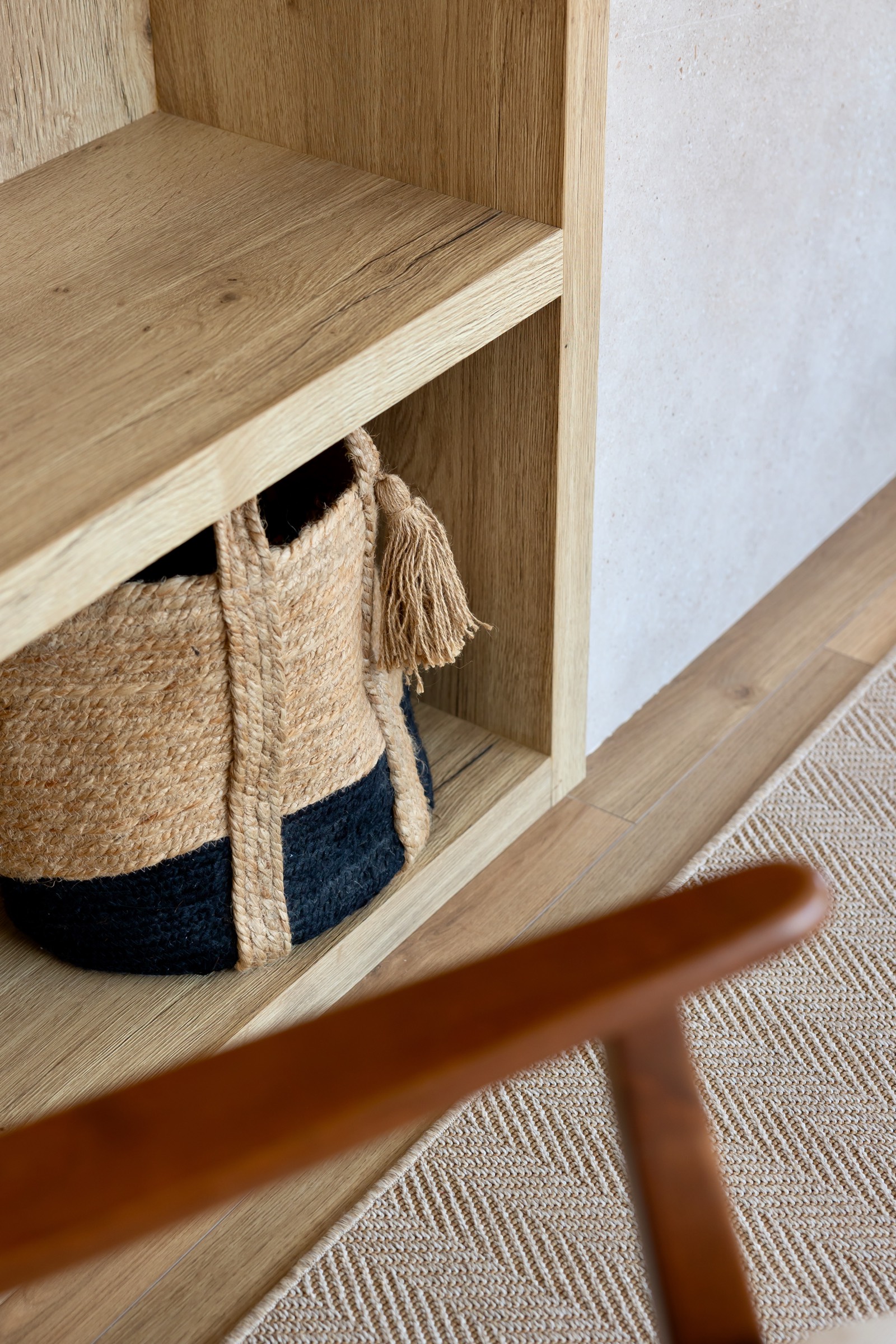 Estantería de madera clara con una cesta de fibra natural y azul oscuro en el estante inferior. Se ve parte de una alfombra beige y la pata de un mueble de madera en primer plano.