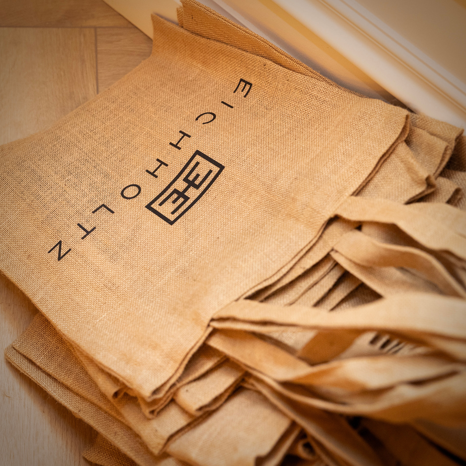 Bolsas de yute apiladas con el logotipo 'E-motion' impreso en negro. Las bolsas son de color beige natural y tienen asas. Están colocadas sobre una superficie de madera clara, posiblemente un suelo.