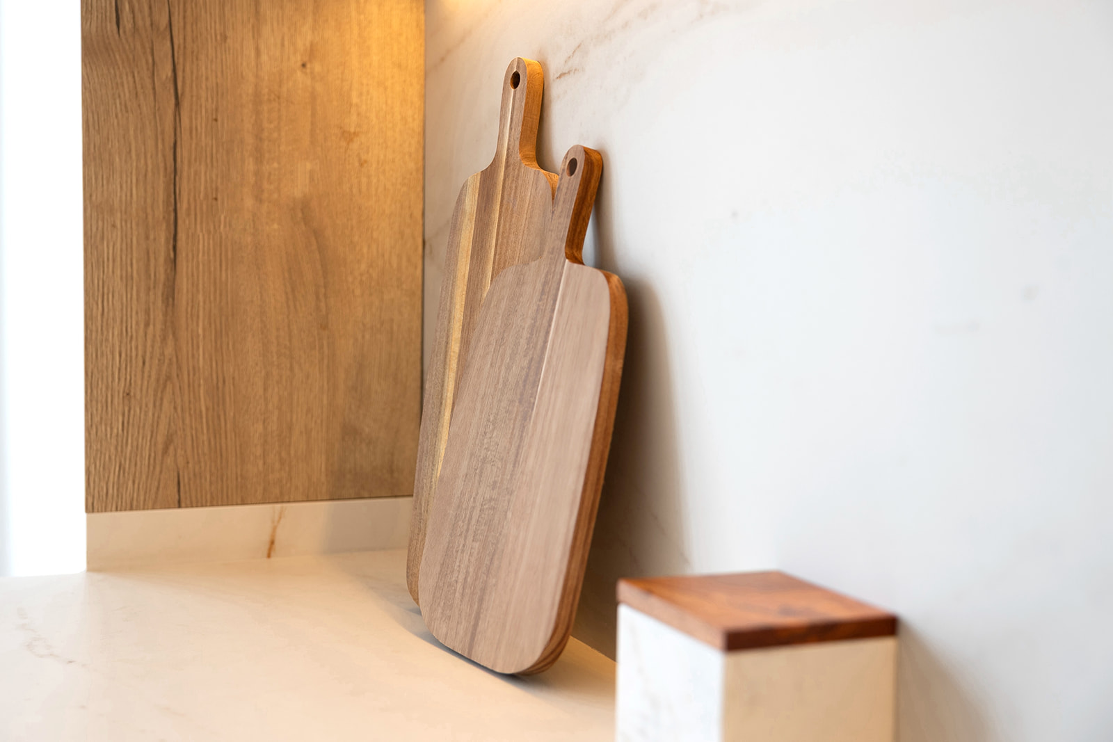 Dos tablas de cortar de madera apoyadas contra una pared blanca en una cocina. Una caja pequeña con tapa de madera se encuentra junto a ellas. La escena muestra un estilo de cocina minimalista y natural.