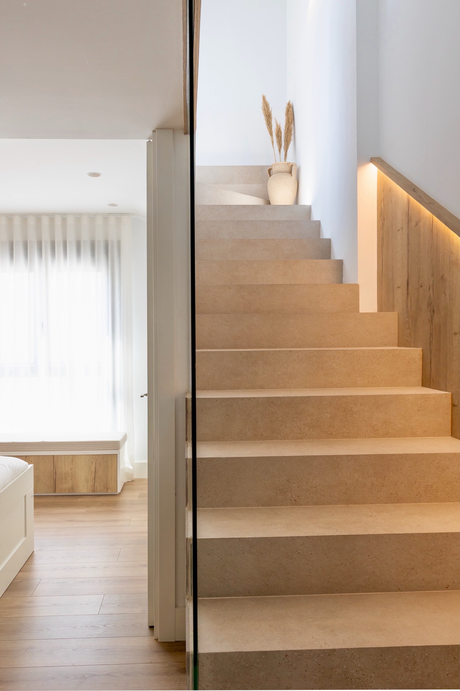 Escalera moderna con peldaños de piedra beige, barandilla de vidrio transparente y decoración con jarrón y hierba seca en el rellano