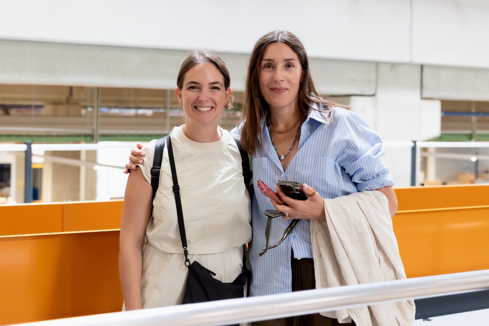 Dos mujeres de mediana edad sonriendo juntas en un espacio de oficina con paredes naranjas y blancas, vestidas con ropa casual profesional
