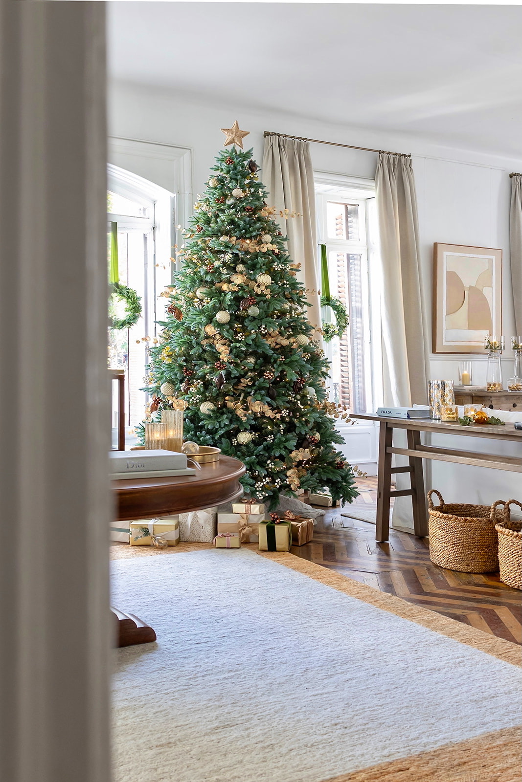 Árbol de Navidad decorado con adornos dorados y blancos en una elegante sala de estar con paredes blancas, cortinas beige, piso de madera en espiga y regalos envueltos debajo del árbol