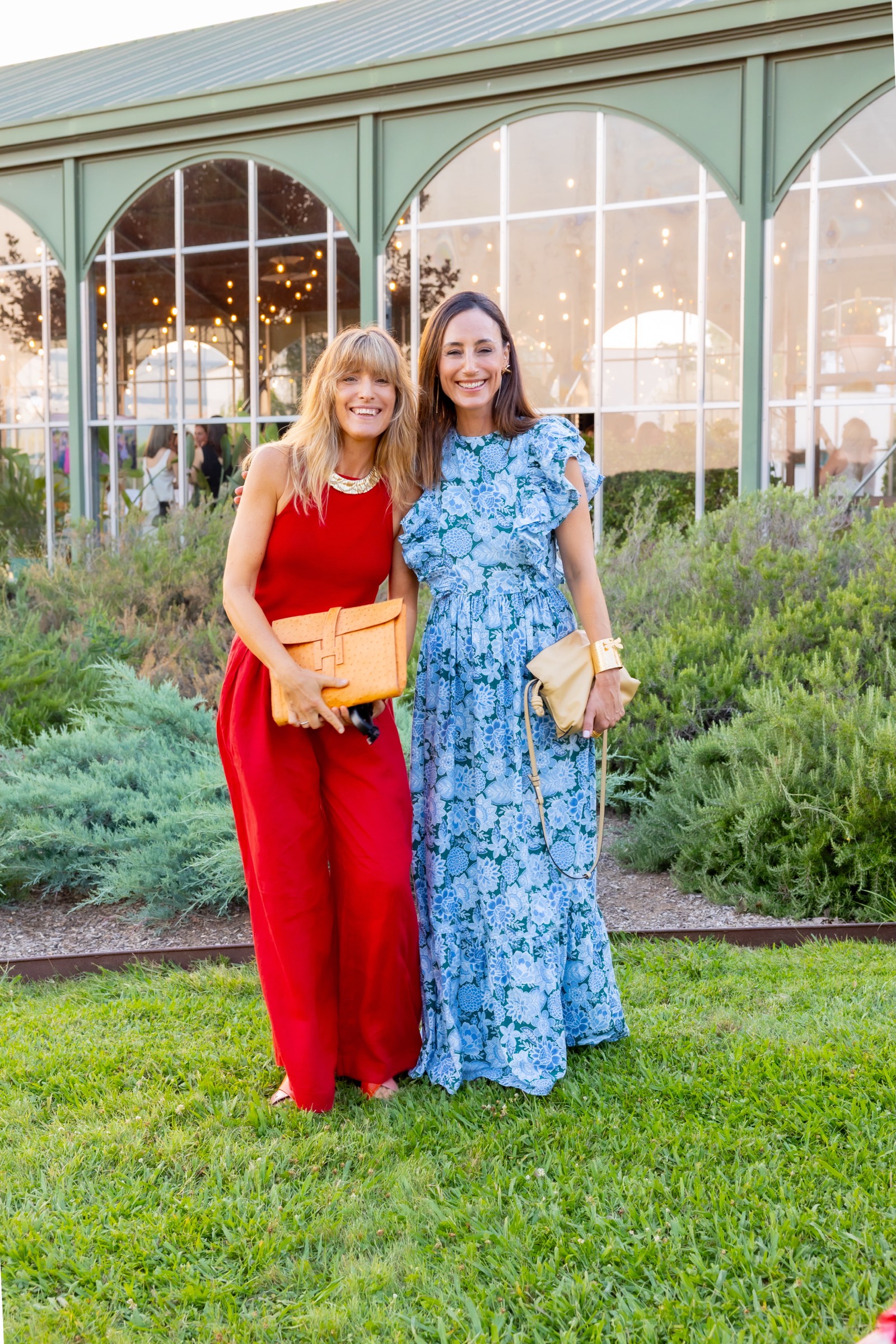 Dos mujeres elegantemente vestidas, una con un vestido rojo largo y otra con un vestido largo estampado en azul, de pie en un jardín con estructura de cristal al fondo