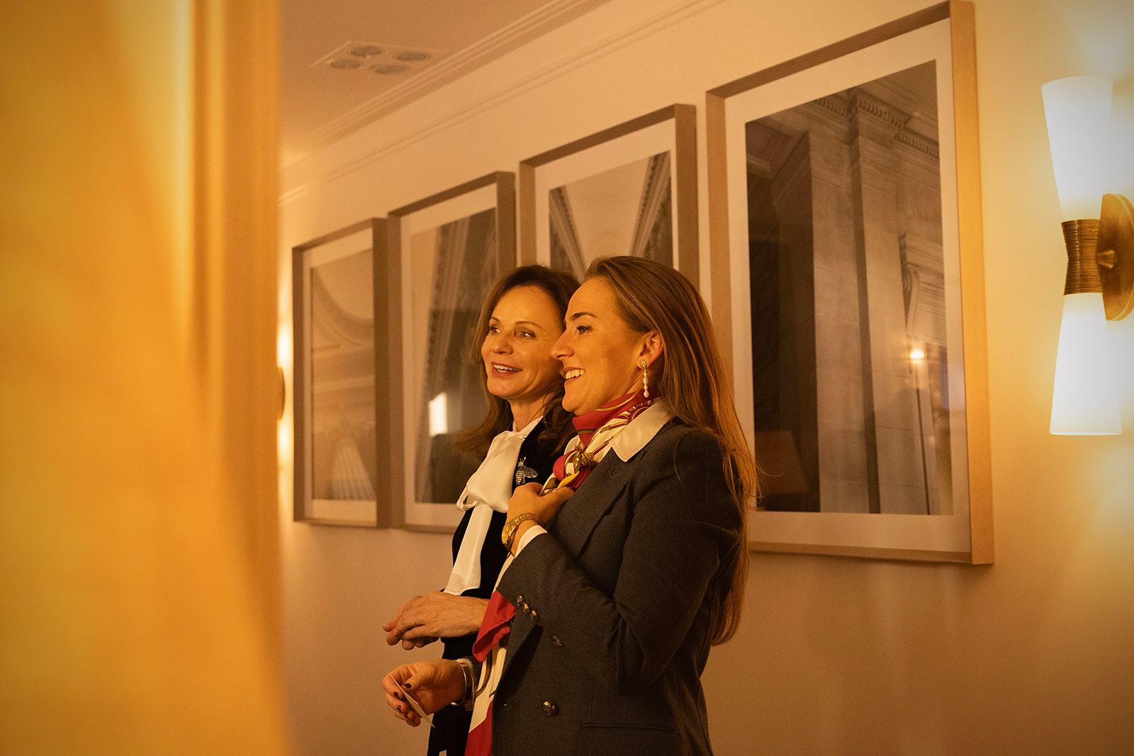 Dos mujeres elegantemente vestidas sonríen en una habitación con iluminación cálida. Detrás de ellas hay cuadros enmarcados en la pared, sugiriendo que están en una galería de arte o espacio similar.