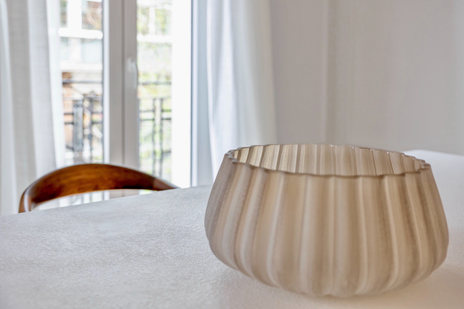 Un cuenco decorativo de vidrio esmerilado con forma ondulada sobre una mesa blanca junto a una silla de madera. Al fondo se ve una ventana con cortinas blancas que deja entrar luz natural.