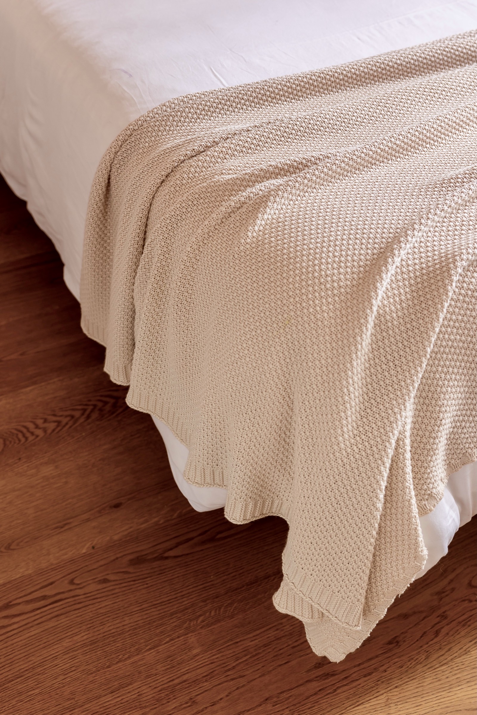 Manta tejida de color beige claro extendida sobre una cama blanca con piso de madera, mostrando textura de punto delicado