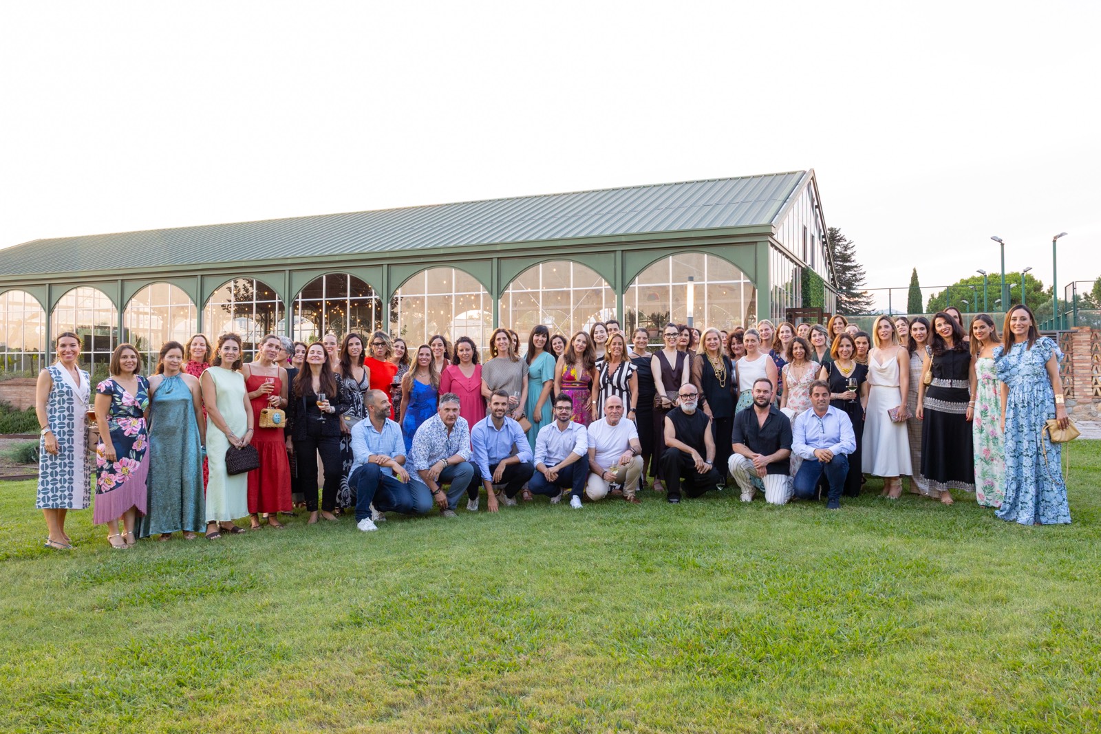 Grupo numeroso de personas vestidas formalmente posando juntas en un césped verde frente a un edificio de cristal con arcos