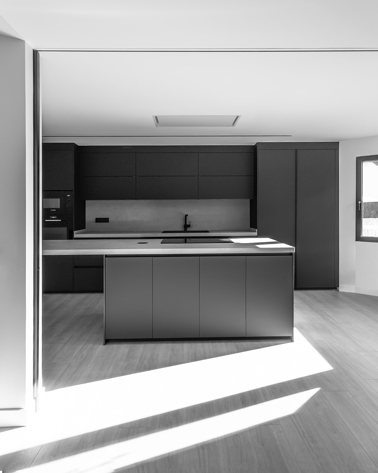 Cocina moderna minimalista en blanco y negro con isla central, armarios oscuros, encimera clara, suelo de madera y luz natural entrando por una ventana, creando un contraste de sombras en el suelo.