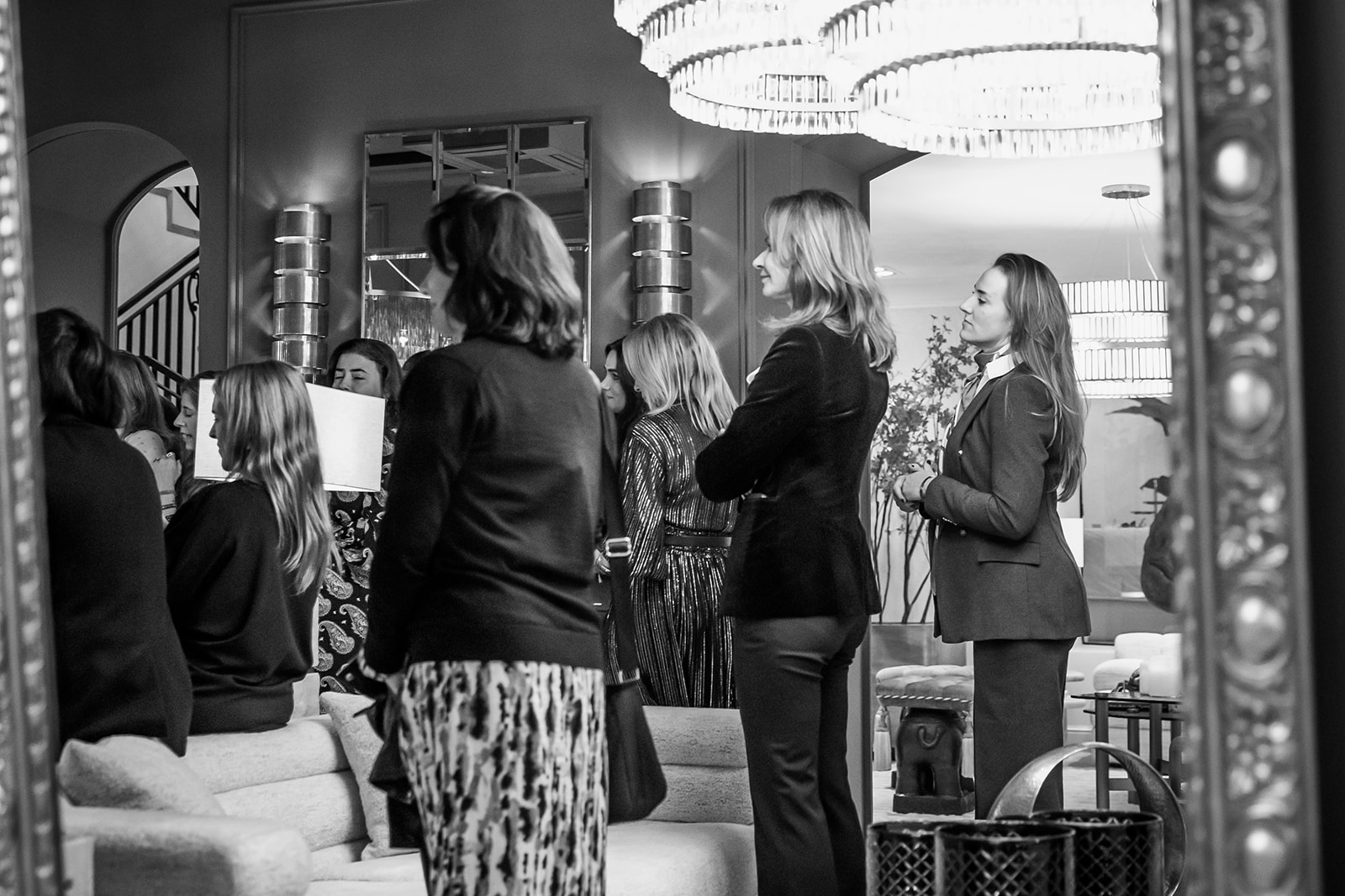 Imagen en blanco y negro de un grupo de personas elegantemente vestidas reunidas en un salón lujoso con una gran lámpara de araña. La escena muestra una atmósfera sofisticada y social.